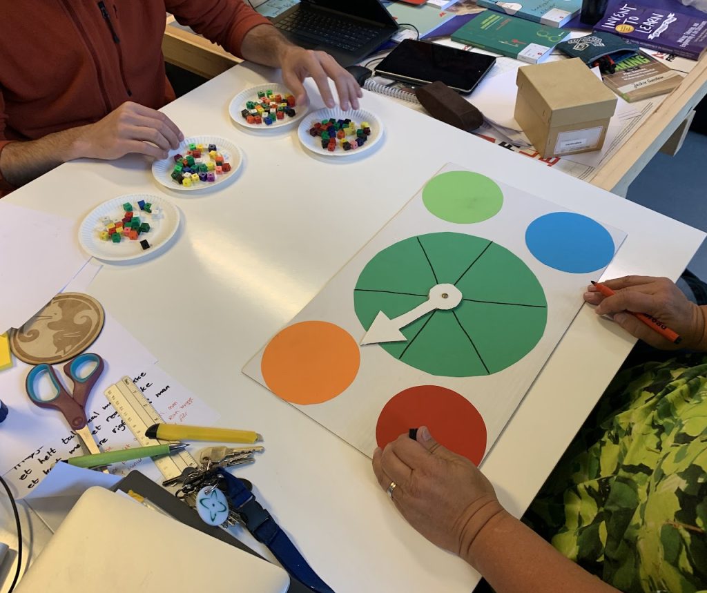 Der afprøves et spil, som er lavet med papir og centicubes. På papiret ses en drejeskive med en pil der kan snurre rundt, og pege på forskellige farver.